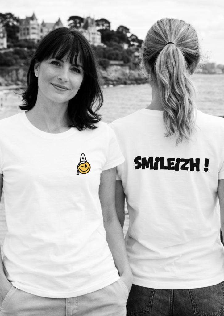 Deux femmes portant un t-shirt avec devant un logo bretonne portant une coiffe traditionnelle avec hermine bretonne et au dos le logo texte smileizh !